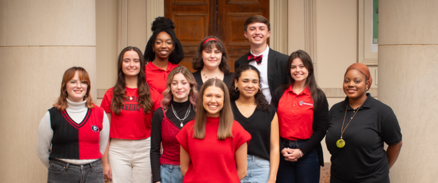 Ten smiling UGA students in red and black, standing on steps in front of the Fine Arts Building columns