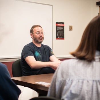 A man talks to two students in the foreground. 